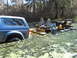 1992 Ford Bronco - A blue 1992 Ford Bronco is partially submerged in a swamp, adjacent to a homemade boat with two seats and an engine.