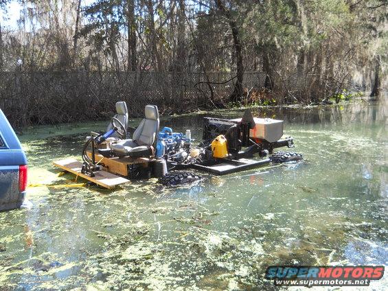 Custom amphibious vehicle with two seats and engine in swampy water. Part of a blue SUV is visible on the left