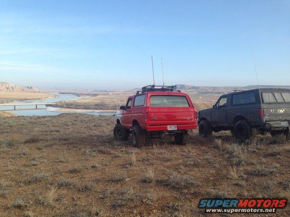 Red 1979 Ford Bronco and a dark SUV parked on a hill with a river in the background