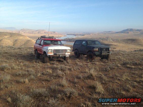 Red and white 1978 Ford Bronco and a dark 1990s Ford Bronco parked on a dry, grassy hillside overlooking a river