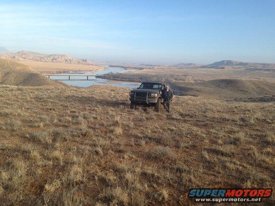 Dark pickup truck on grassy hill with a person, river and bridge in background