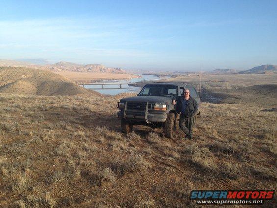 Man leaning on a dark SUV parked on a dry, grassy hill overlooking a river and bridge