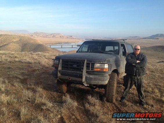 Man standing next to a dark-colored Ford Bronco in a dry, hilly landscape with a bridge and water in the background