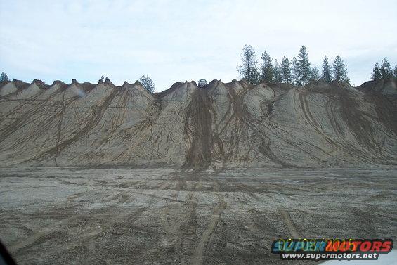 SUV driving up a steep, rutted dirt hill with tire tracks, trees on the horizon