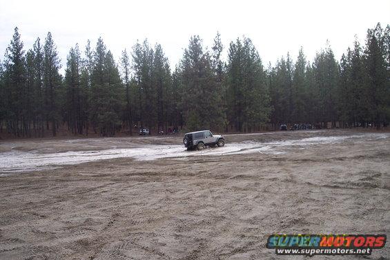 Silver Jeep Wrangler driving on a dirt track with trees in the background. Other vehicles are visible further back