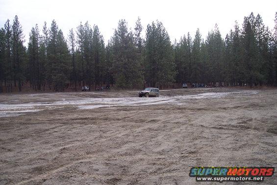 Jeep Wrangler driving through mud with trees in background. Other vehicles and people are visible further back