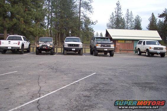 Five SUVs and trucks parked in a lot, including a white Ford F-150, a grey Jeep Wrangler, a white Ford Bronco, and two other Ford Broncos. A building and trees are in the background