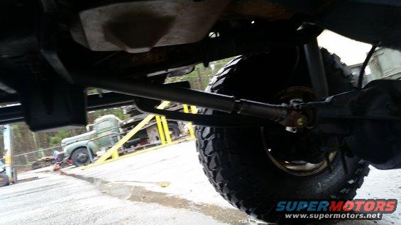 Underside view of a vehicle's drivetrain and tire, with an old truck on a flatbed trailer in the background