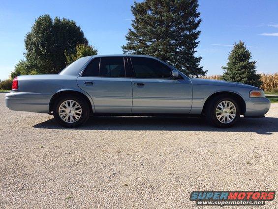 Side view of a 2003 Ford Crown Victoria parked on a gravel lot with trees and a cornfield in the background