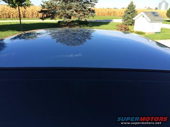 Dark blue car roof reflecting trees, cornfield, and a white shed under a blue sky