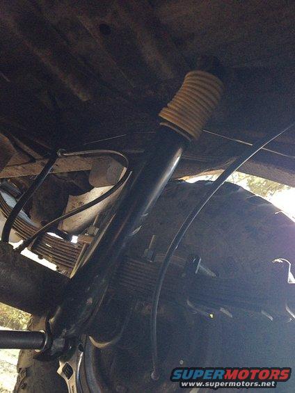 Undercarriage of a vehicle showing a shock absorber with a yellow boot, leaf springs, and a tire. Text 'SUPERMOTORS' is visible
