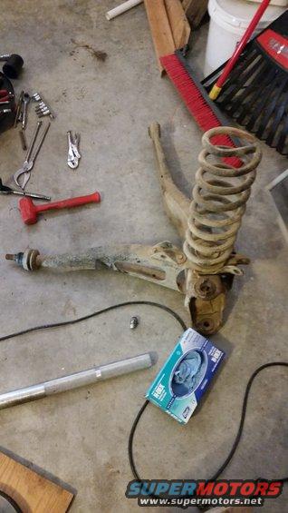 Disassembled front suspension parts, including a coil spring and control arm, lie on a concrete floor next to tools and a box of gloves