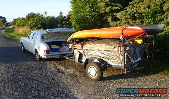 white sedan with open trunk towing a trailer loaded with kayaks and camping gear