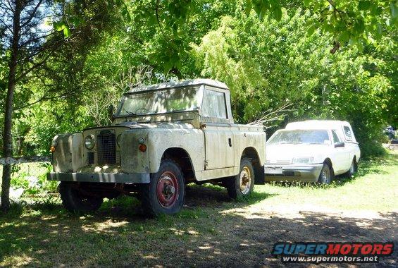Weathered Land Rover pickup truck parked next to a white sedan in a grassy area with trees