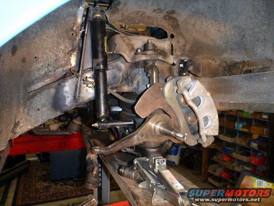 Car's front suspension with shock, brake caliper, and control arms visible. A floor jack is under the frame