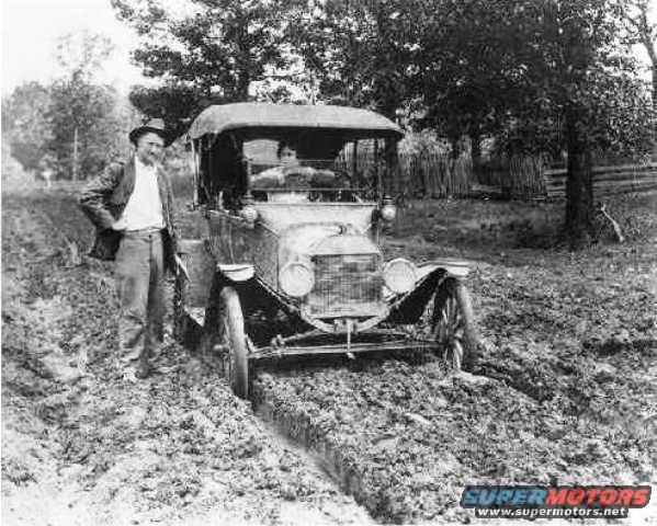 Man in hat stands next to a 1915 Ford Model T stuck in a muddy rut. Another person is in the driver's seat