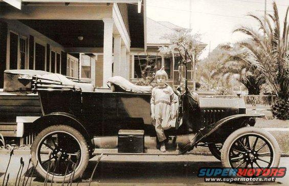 Boy in a light-colored outfit stands on the running board of a dark-colored touring car. A house with a porch is behind them