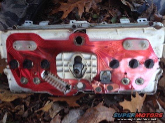 Dashboard instrument cluster with red circuit board and black circular gauge openings, lying on fallen leaves