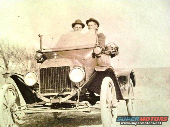 Two men in hats ride in an early automobile. The car has a tall grille and large round headlights