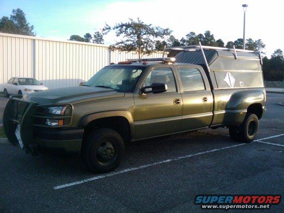 Olive green dually pickup truck with a utility cap and brush guard parked in a lot. White sedan in background