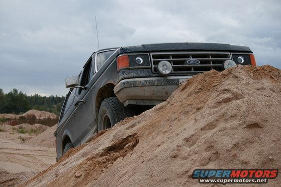 Black 1987 Ford Bronco driving up a large sand dune with a cloudy sky above