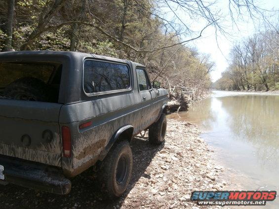 Muddy 1978 Ford Bronco parked on rocky shore next to a river
