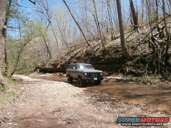 Gray Ford Bronco driving through shallow water on a dirt path in a wooded area