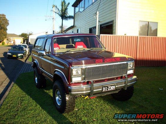 Maroon Ford Bronco with lifted suspension and aftermarket grille. Two other cars are parked on the street