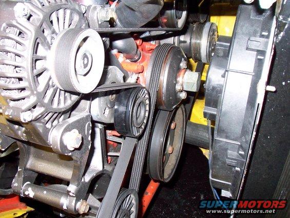 Close-up of an engine's alternator, pulleys, and belts. A black electric fan is visible on the right