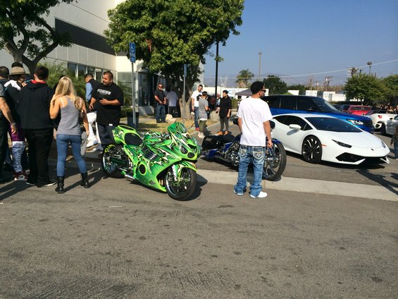 Two custom motorcycles and a white Lamborghini sports car parked on a street with people around