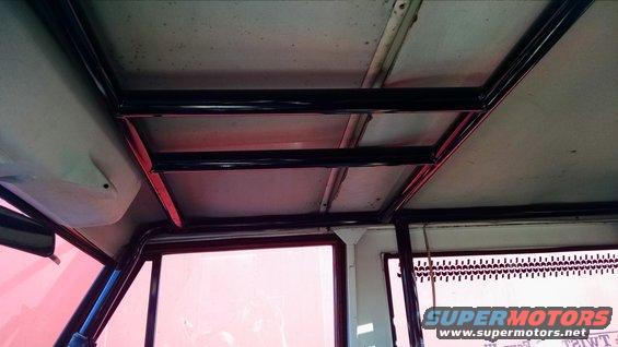 Interior view of a vehicle's roof with a black metal roll cage and red trim. Pinkish light filters through the windows