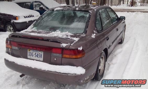 Brown 1996 Subaru Legacy sedan parked in snow, rear view. Another vehicle covered in snow is to the left