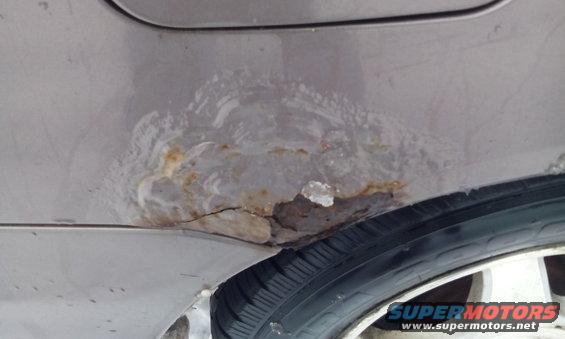 Close-up of a heavily rusted and cracked wheel well on a light-colored vehicle, with a tire visible