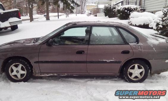 Brown sedan in snow, side view. Snow covers the vehicle and the ground. A dark pickup truck is partially visible on the left