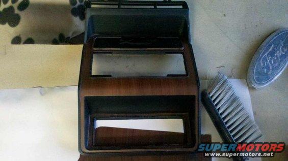 Car dashboard trim with wood grain and two rectangular openings, next to a brush and a Ford emblem