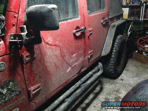 Side view of a red Jeep Wrangler with aftermarket side steps and a soft top. It's dirty and parked in a garage