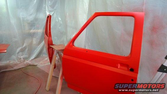 Red car door and fender on sawhorses, being painted. A Supermotors.net watermark is visible