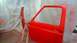 1983 Ford Bronco - A red car door and fender are being painted in a workshop. The parts are mounted on sawhorses.