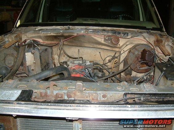 Engine bay of a Ford truck, showing carburetor, brake master cylinder, and wiring. Hood latches are visible in the foreground