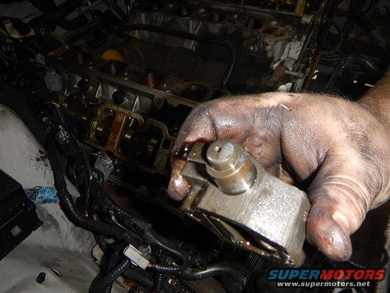 Dirty hand holding a metal engine component over an open engine block with visible wiring