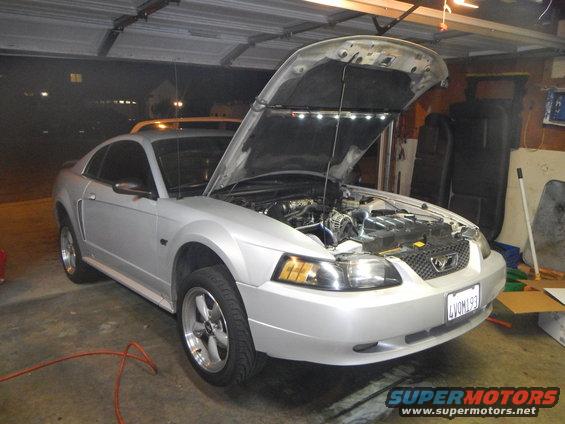 Silver 2002 Ford Mustang coupe with hood open in a garage, showing the engine bay. Lights are installed under the hood