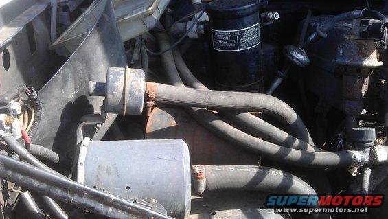 Engine bay with hoses, a black cylinder with a label, and metal components. Some rust is visible