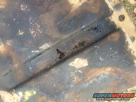 Underside of a metal part, heavily stained with oil and grime, with a hole and some debris