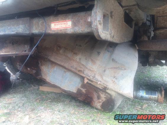 Vehicle underbody showing a rusty fuel tank and frame with a warning sticker