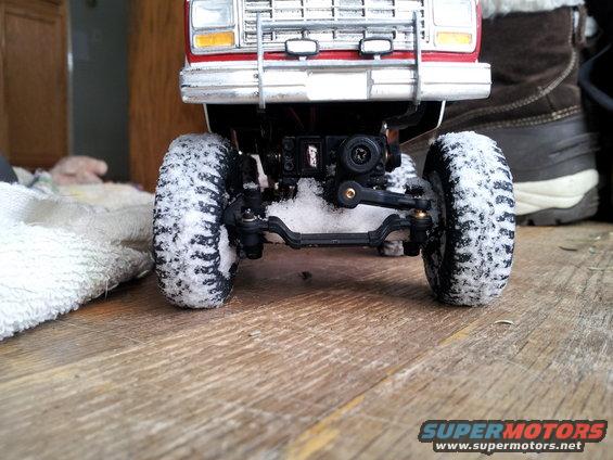 Toy truck front axle and wheels covered in snow, with a red body and chrome grille