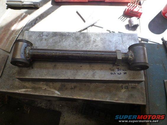 Custom suspension control arm with rubber bushings and a nut on a threaded end, resting on a metal surface with handwritten notes