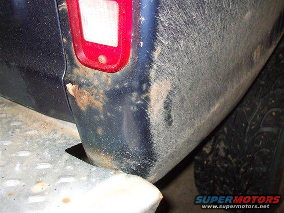 Rear quarter panel of a dark blue vehicle showing rust damage near the taillight and mud splatter