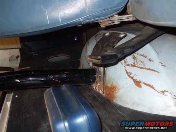 Close-up of vehicle interior floor area under the seat, showing rust and a black metal bar