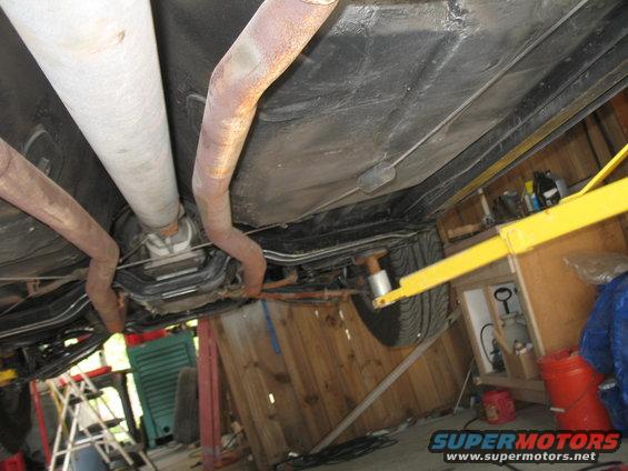 Underside of a vehicle on a lift, showing exhaust pipes, drive shaft, and a tire