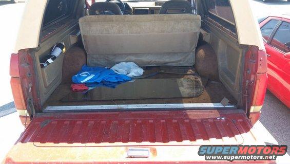 Red and tan Ford Bronco cargo area with folded rear seat and items on the floor. A red car is parked to the right